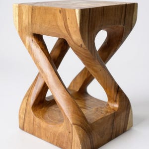 May include: A handcrafted wooden stool with a unique hourglass-like design. The stool is made of light brown wood and features a square top and base, with curved, intersecting supports. The open design adds a modern touch to this functional piece.