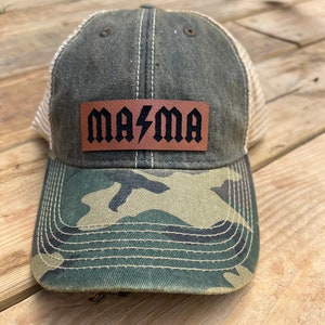 May include: A green and brown camouflage trucker hat with a brown leather patch that says "MA/MA" in black letters with a lightning bolt between the words.