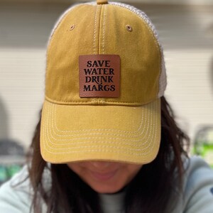 May include: A yellow and white trucker hat with a brown leather patch that reads "SAVE WATER DRINK MARGS".