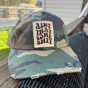 May include: Camouflage baseball cap with distressed edges. A rectangular patch on the front displays the phrase "AINT THAT SOME SHIT" in a stacked, stylized font. The cap is resting on a wicker surface.