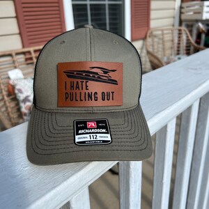 May include: A green and black trucker hat with a brown leather patch that says "I hate pulling out". The patch features a silhouette of a boat. The hat is adjustable and has a Richardson 112 label.