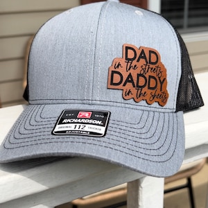 May include: A gray and black trucker hat with a brown leather patch that reads "Dad in the streets, Daddy in the sheets". The hat is adjustable and has a Richardson 112 label.