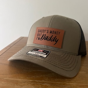 May include: A green and black trucker hat with a brown leather patch that says "Daddy's Money But I'm Daddy". The hat is made by Richardson 112.