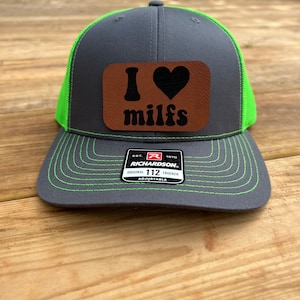 May include: A gray and green trucker hat with a brown leather patch that says "I love milfs". The hat is adjustable and has the Richardson logo on the back.