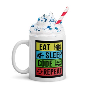 May include: A white ceramic mug featuring a colorful graphic with the text EAT, SLEEP, CODE, REPEAT, accompanied by icons of a plate, a sleeping person, a computer monitor with code brackets, and a refresh symbol, topped with white whipped cream.