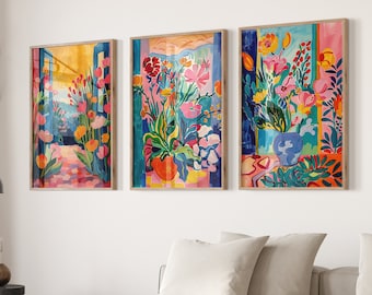May include: Three colorful abstract paintings of floral arrangements in a modern living room setting. The paintings feature vibrant colors, including pink, yellow, blue, and green, and depict stylized flowers and foliage. The paintings are framed in natural wood and are hung on a white wall.