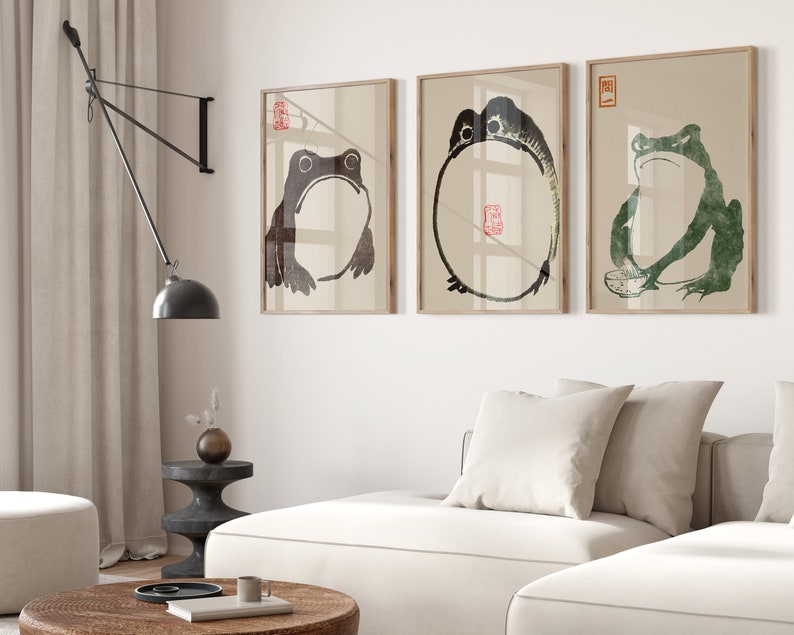 May include: Three framed black and white ink drawings of frogs. The drawings are on a white wall above a white couch with a white pillow. A black lamp with a black shade is in the corner of the room.