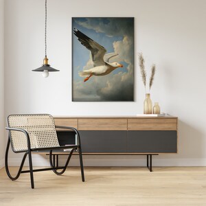 Puede incluir: Una pintura enmarcada de una gaviota blanca en vuelo contra un cielo azul con nubes blancas. La pintura est&aacute; colgada en una pared blanca sobre un gabinete de madera con cajones negros.