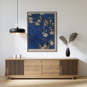 May include: A vintage Japanese woodblock print of a bird perched on a branch of cherry blossoms. The print is framed and hanging on a white wall above a wooden cabinet.