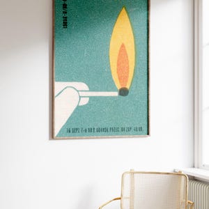 May include: A framed vintage-style poster featuring a match with a burning flame. The flame is yellow and orange against a teal background. The poster includes text at the top and bottom. The frame is a dark brown color.