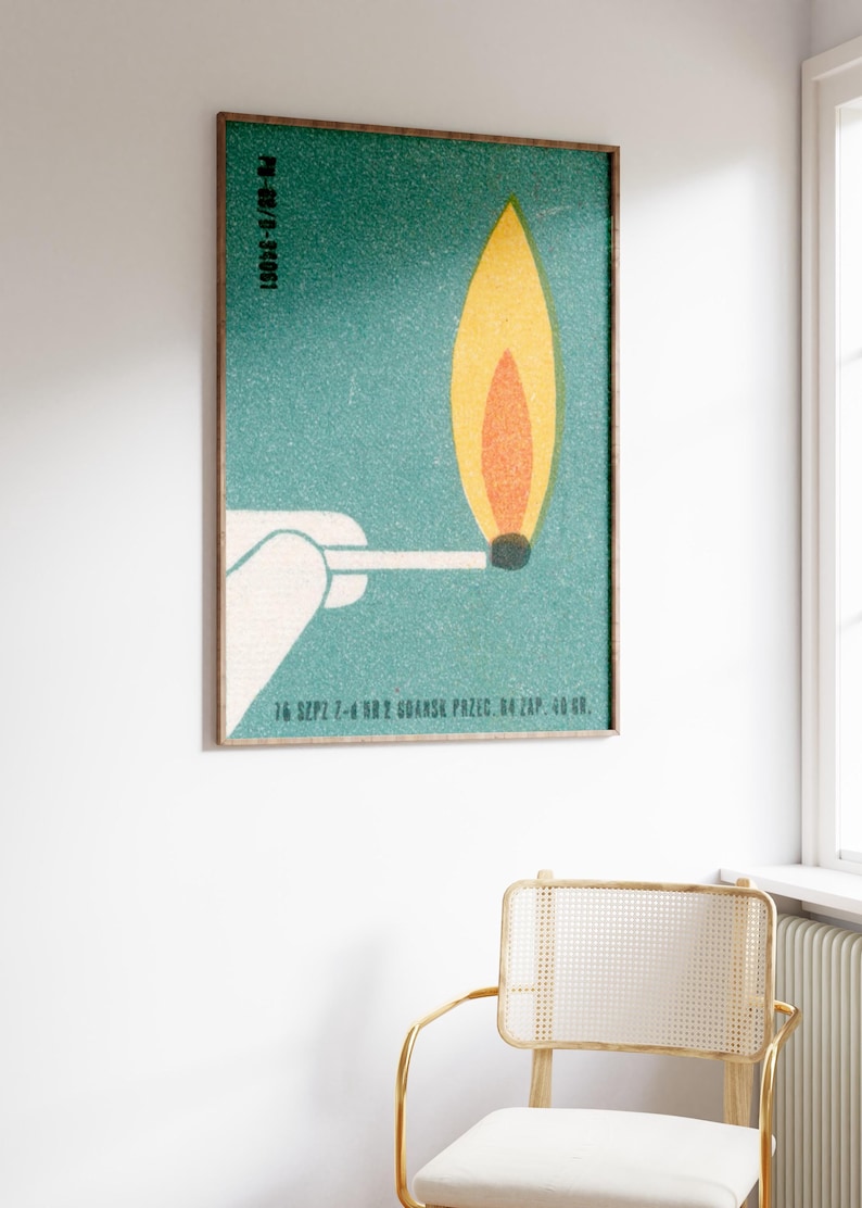 May include: A framed vintage-style poster featuring a match with a burning flame. The flame is yellow and orange against a teal background. The poster includes text at the top and bottom. The frame is a dark brown color.