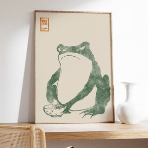 May include: A framed print of a green frog sitting with a bowl of food. The frog has a serious expression on its face. The print has a Japanese character in the top left corner.