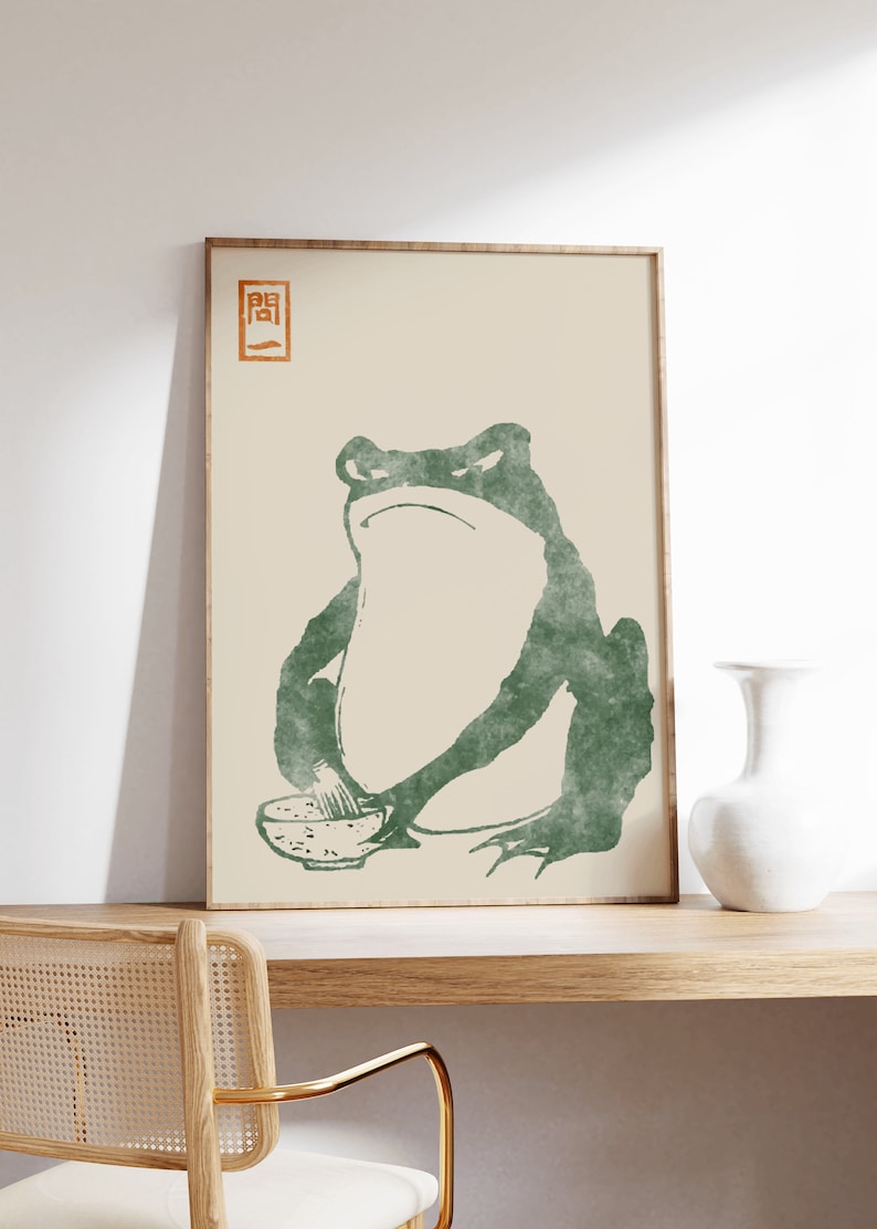 May include: A framed print of a green frog sitting with a bowl of food. The frog has a serious expression on its face. The print has a Japanese character in the top left corner.