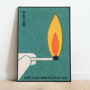May include: A framed poster featuring a stylized illustration of a lit match. The flame is depicted in shades of yellow and orange against a teal background. The match is held by a white hand. Text is visible at the bottom.