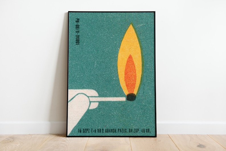 May include: A framed poster featuring a stylized illustration of a lit match. The flame is depicted in shades of yellow and orange against a teal background. The match is held by a white hand. Text is visible at the bottom.
