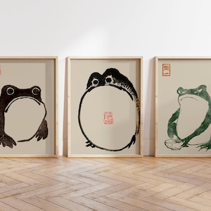 May include: Three framed prints of black and green frogs on a beige background. The frogs are in different poses, one is sitting, one is standing, and one is in a circle shape. Each print has a red or green stamp in the top corner.