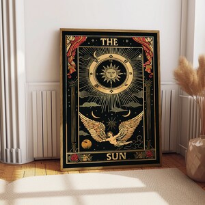 Op de afbeelding: De tarotkaart De Zon heeft een zwarte achtergrond met gouden accenten. Een ge Fl&uuml;gelde engel vliegt door de lucht met een zon en sterren op de achtergrond. De kaart heeft bovenaan "THE SUN" en onderaan "SUN" staan.