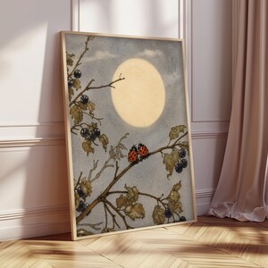 May include: A framed art print showing a watercolour illustration of a full moon in a cloudy sky. A tree branch with leaves, black berries, and two ladybirds is in the foreground. The artwork is in a wooden frame and leans against a wall.