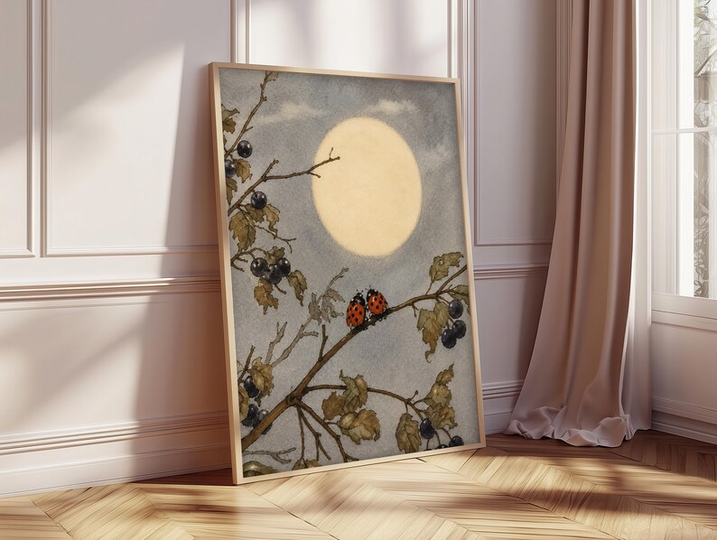 May include: A framed art print showing a watercolour illustration of a full moon in a cloudy sky. A tree branch with leaves, black berries, and two ladybirds is in the foreground. The artwork is in a wooden frame and leans against a wall.