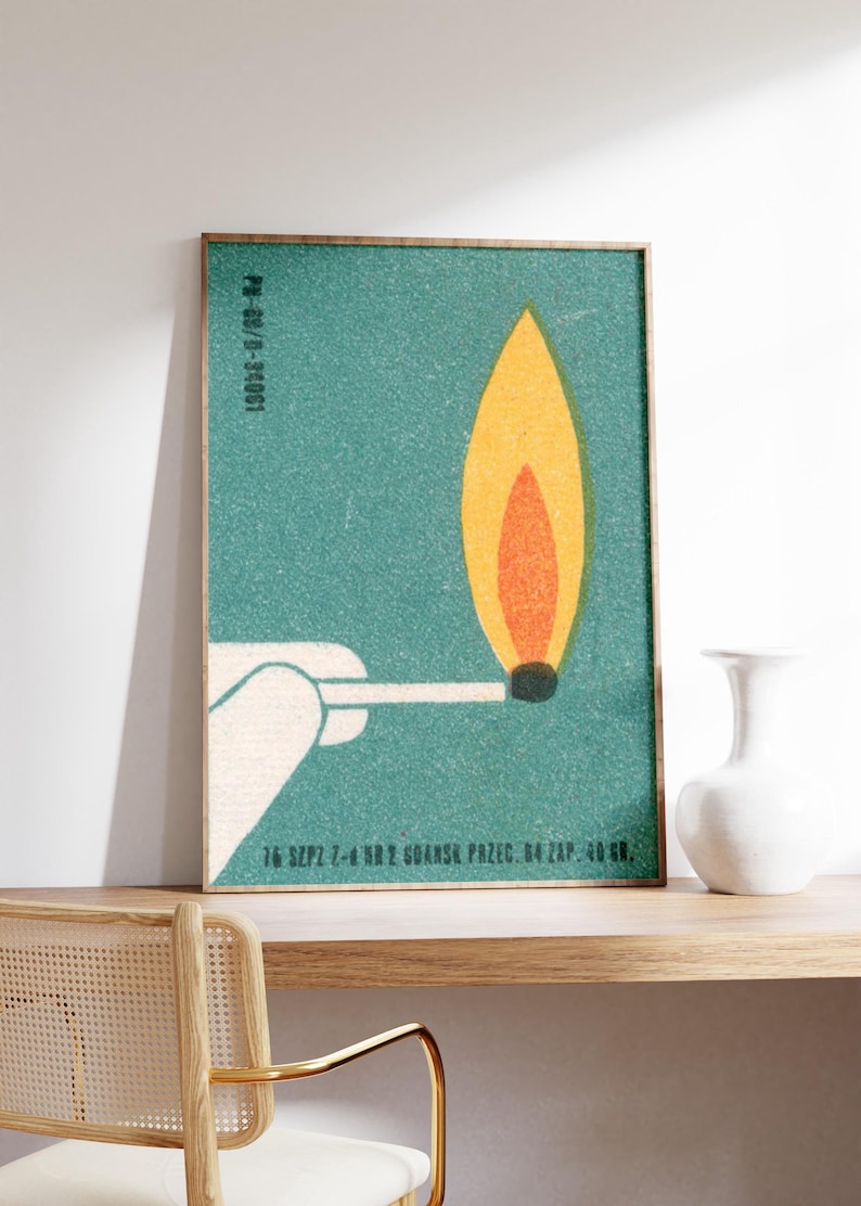 May include: A framed art print featuring a stylized match with a burning flame. The flame is yellow and orange against a teal background. The match is held by a white hand. Text at the top and bottom of the print.