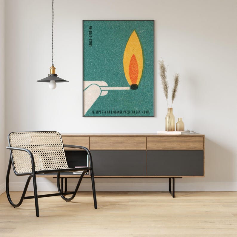 May include: A framed teal poster with a match and flame illustration. The flame is yellow and orange. The poster is hanging above a wooden cabinet with black drawers. A black chair with a woven seat is in the foreground.