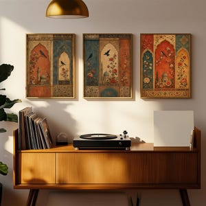 May include: Three framed wall art pieces with ornate, vintage-style designs. Each features a central archway motif with birds, flowers, and architectural elements in shades of red, blue, and gold. The artwork hangs above a wooden cabinet with a record player.