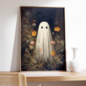 K&ouml;nnte beinhalten: Ein gerahmter Kunstdruck mit einem wei&szlig;en Geist mit schwarzen Augen in einem Blumenfeld. Das Kunstwerk hat eine Vintage-&Auml;sthetik mit dunklem Hintergrund und Holzrahmen. Die Blumen sind orange und wei&szlig;.