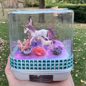 May include: A clear, rectangular terrarium with a white base, decorated with a teal gem-studded band. Inside, a miniature scene features two unicorn figurines, pink and purple flowers, and a large pink rose. The background is a purple and white galaxy design.
