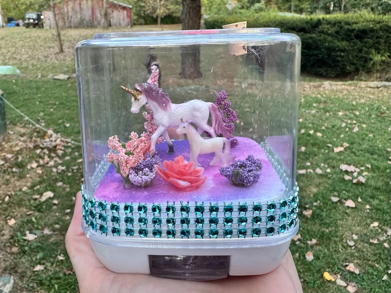 May include: A clear, rectangular terrarium with a white base, decorated with a teal gem-studded band. Inside, a miniature scene features two unicorn figurines, pink and purple flowers, and a large pink rose. The background is a purple and white galaxy design.