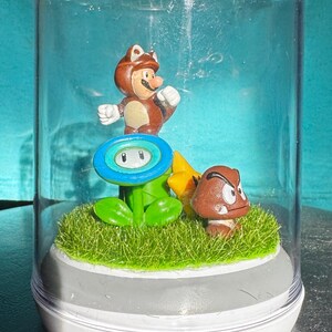 May include: A clear glass dome encases a miniature Super Mario diorama. The scene features a brown figure with a cat-like hat, a blue flower, a yellow star, and a brown Goomba on a bed of green faux grass, all set on a white base.