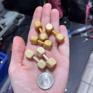 May include: A hand holding two sets of miniature wooden dumbbells. The dumbbells are made of light and dark wood. The smaller set is light wood and the larger set is dark wood.