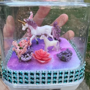 May include: A clear plastic container decorated with teal rhinestones, housing a miniature fantasy scene. Inside, two white unicorn figurines with purple manes stand on a purple and pink base, surrounded by pink and purple floral accents.