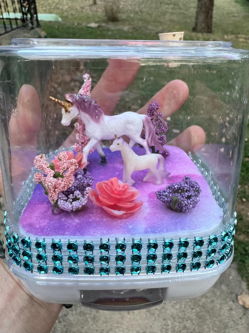 May include: A clear plastic container decorated with teal rhinestones, housing a miniature fantasy scene. Inside, two white unicorn figurines with purple manes stand on a purple and pink base, surrounded by pink and purple floral accents.