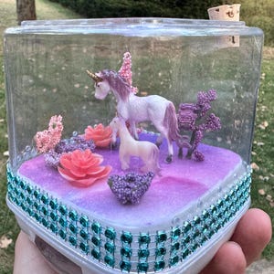 May include: A clear, rectangular terrarium with a decorative base, featuring two unicorn figurines, pink flowers, and purple accents. The base is trimmed with teal-colored jewels. The terrarium is held in a person's hand.