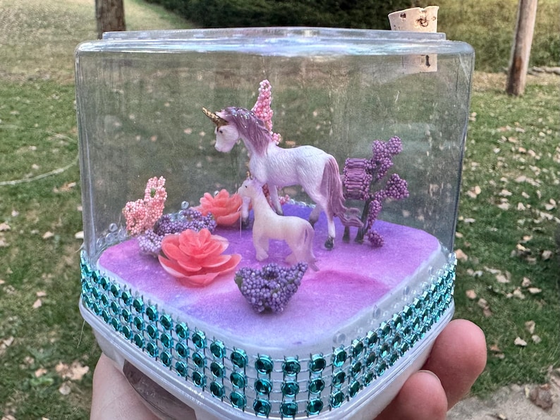 May include: A clear, rectangular terrarium with a decorative base, featuring two unicorn figurines, pink flowers, and purple accents. The base is trimmed with teal-colored jewels. The terrarium is held in a person's hand.