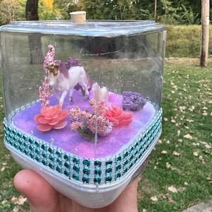 May include: A clear, square terrarium with a white unicorn figurine, pink and coral flowers, and purple accents. The base is decorated with teal gems. A cork stopper is on top.