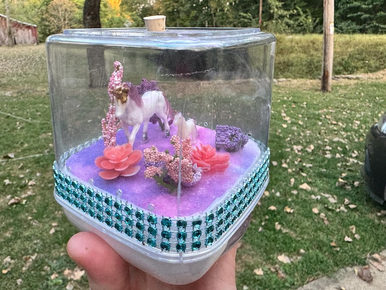 May include: A clear, square terrarium with a white unicorn figurine, pink and coral flowers, and purple accents. The base is decorated with teal gems. A cork stopper is on top.