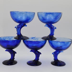 Könnte beinhalten: Fünf blaue Champagner-Coupe-Gläser. Jedes Glas hat einen delfinförmigen Stiel und eine runde Schale. Das Blau hat einen marmorierten Effekt. Die Gläser sind in einer gestapelten Formation angeordnet.