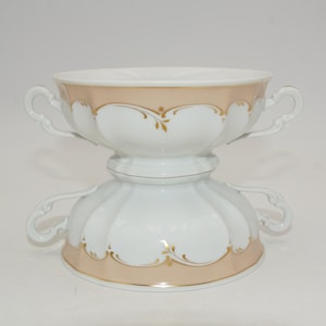 Puede incluir: Dos cuencos de sopa de porcelana blanca apilados, con una banda color melocotón y detalles florales dorados. Cada cuenco tiene dos asas ornamentadas. Los cuencos están diseñados con un borde festoneado.