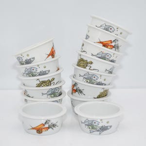 Può includere: Una pila di ciotole in ceramica bianca con una varietà di disegni colorati di pesci, tartarughe e carote. Le ciotole sono tutte della stessa dimensione e forma, e sono impilate l'una sull'altra.