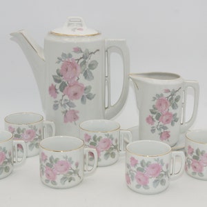 Può includere: Un set da tè in porcellana bianca con un motivo floreale. Il set comprende una teiera, un creamer e sei tazze e piattini. Il motivo floreale presenta rose rosa e foglie verdi.