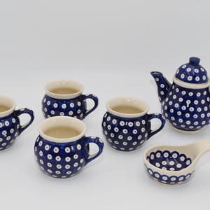 Może przedstawiać: Ceramiczny zestaw do herbaty składający się z czajnika z pokrywką, czterech zaokrąglonych kubków i małej podstawki pod łyżeczkę. Każdy element ozdobiony jest ciemnoniebieskim szkliwem w powtarzający się wzór białych kółek z niebieskim środkiem.