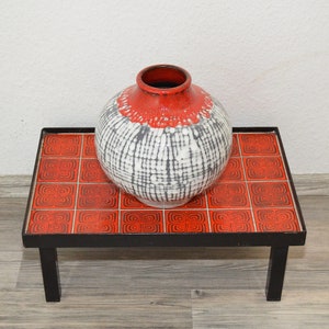 Op de afbeelding: Een zwarte metalen bijzettafel met een rood en zwart betegeld blad. Een witte en grijze keramische vaas met een rode rand staat op de tafel.