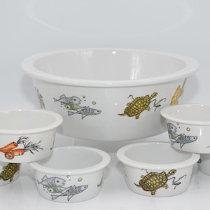 Può includere: Un set di ciotole in ceramica bianca con un design fantasioso che presenta pesci, tartarughe e carote. Le ciotole sono di varie dimensioni, con la ciotola più grande che è una ciotola da portata.
