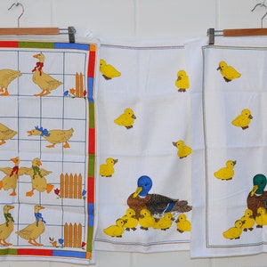 May include: Three white tea towels with different designs. The first towel has a grid pattern with yellow ducks and fences. The second towel has a yellow duck design. The third towel has a yellow duck design with a larger duck and ducklings.