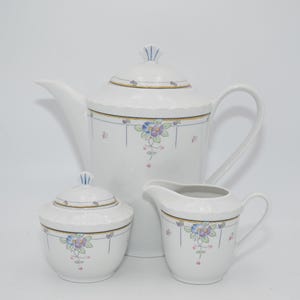 Op de afbeelding: Een wit porseleinen theeservies met een bloemenmotief. De set bevat een theepot, een roomkan en een suikerpot. De bloemen zijn blauw, roze en geel.