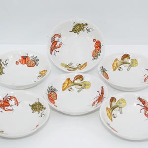 Può includere: Set di sei ciotole in ceramica bianca con disegni colorati di aragoste, tartarughe, funghi e pomodori. Le ciotole sono perfette per servire spuntini o dessert.