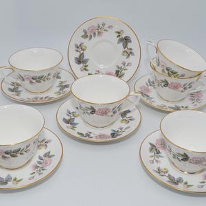 Könnte beinhalten: Ein weißes Teeservice mit Tassen und Untertassen, verziert mit einem floralen Muster aus rosa Rosen und grünen Blättern. Das Set umfasst sechs Tassen, sechs Untertassen und einen Servierteller, alle mit Goldrand.