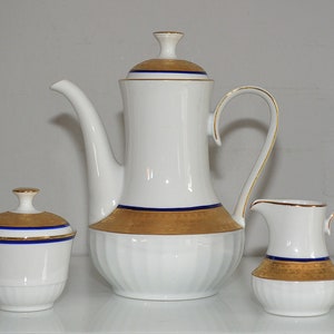 Op de afbeelding: Witte porseleinen koffiepot, roomkan en suikerpot met gouden en blauwe versiering. De set heeft een klassiek ontwerp met een geribbeld lichaam.