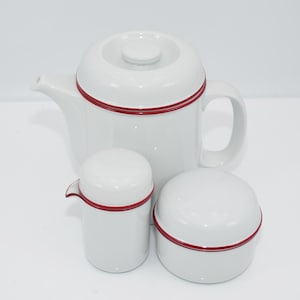Könnte beinhalten: Ein weißes Tee-Set bestehend aus Teekanne, Milchkännchen und Zuckerdose mit einem roten Streifen um jedes Stück.